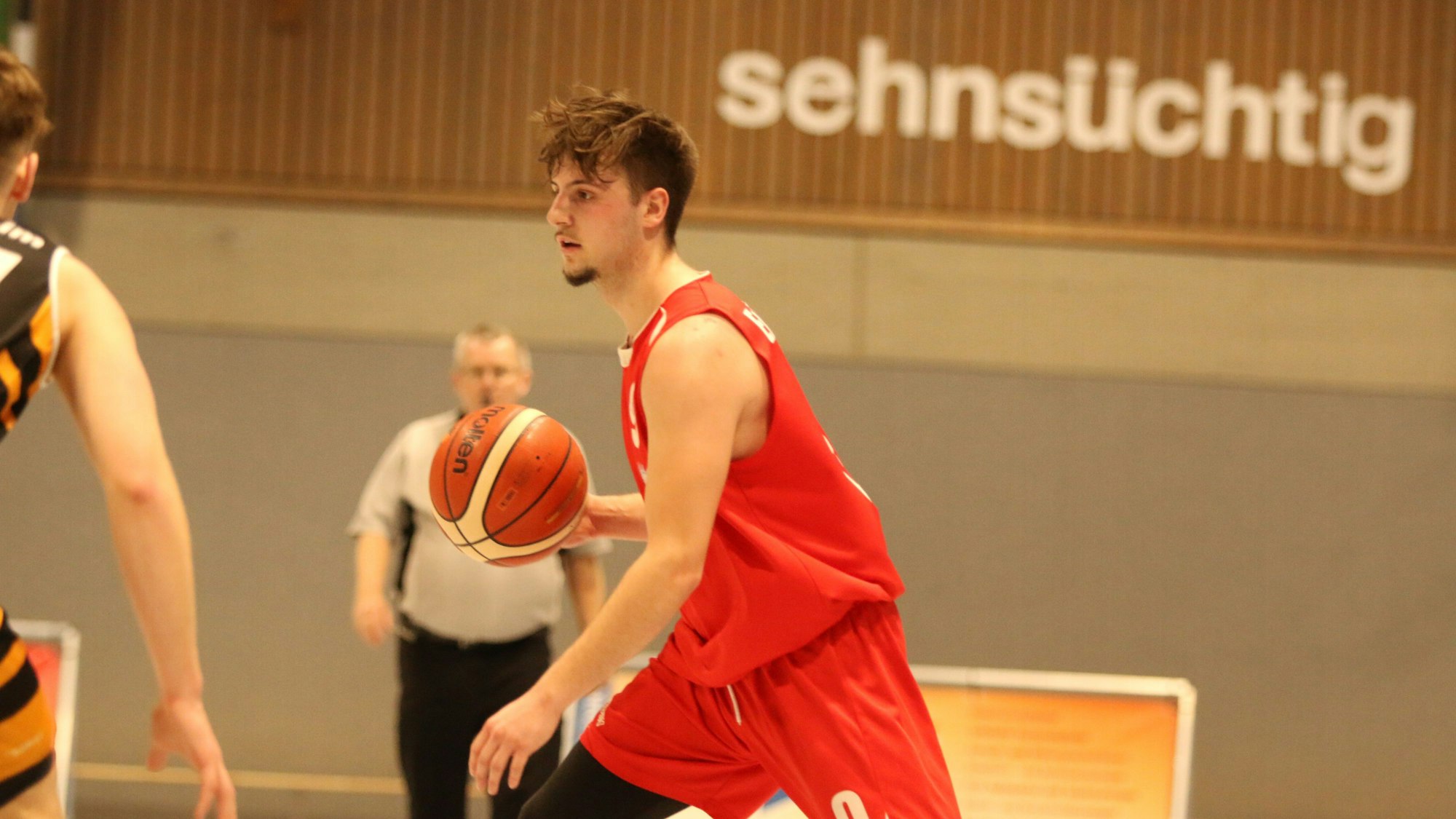 Aufbauspieler Jan Züll dribbelt mit dem Ball. Im Hintergrund steht an der Wand der Sporthalle das Wort „sehnsüchtig“.