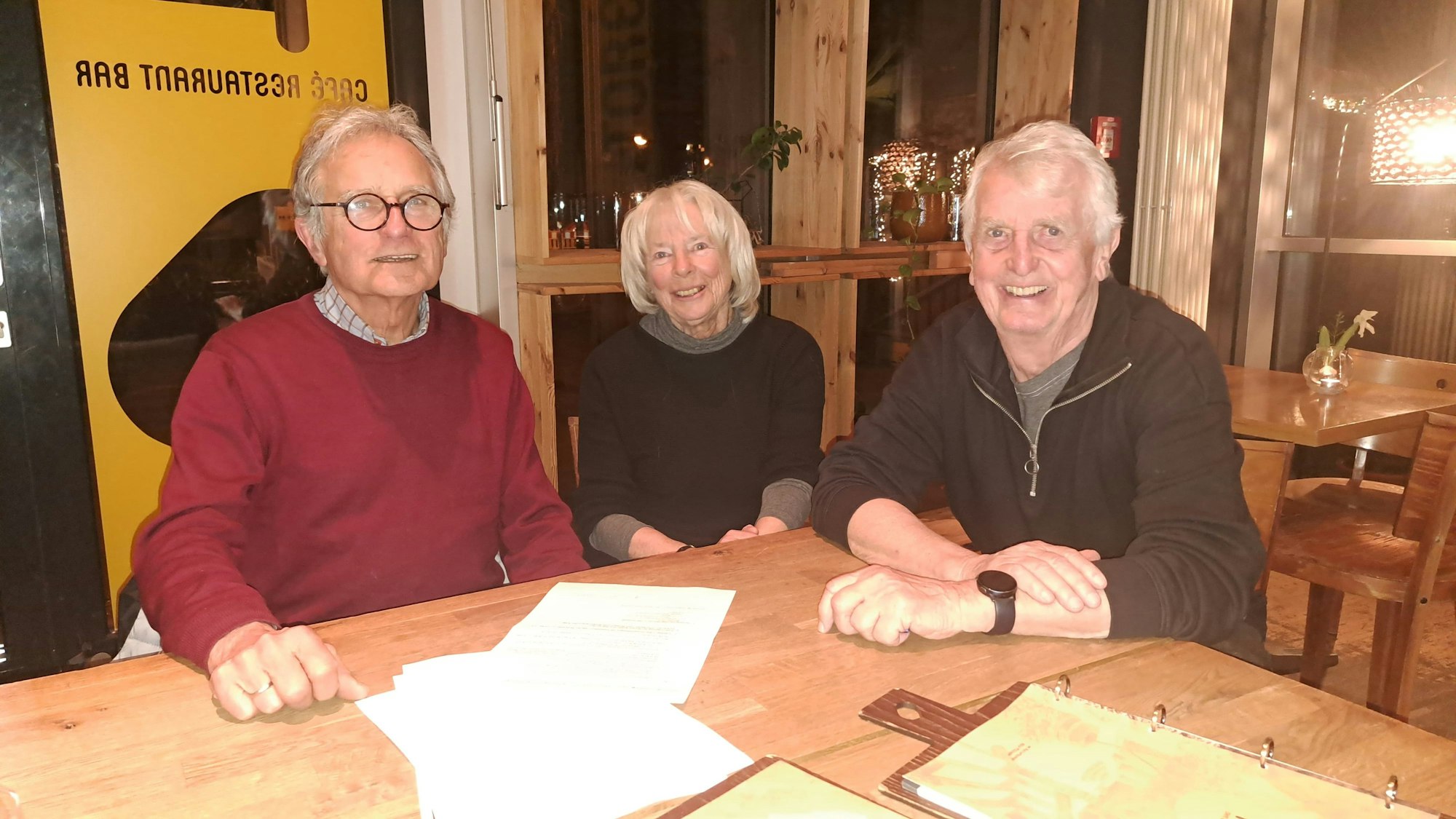 Zwei ältere Männer und eine Frau sitzen an einem Holztisch.
