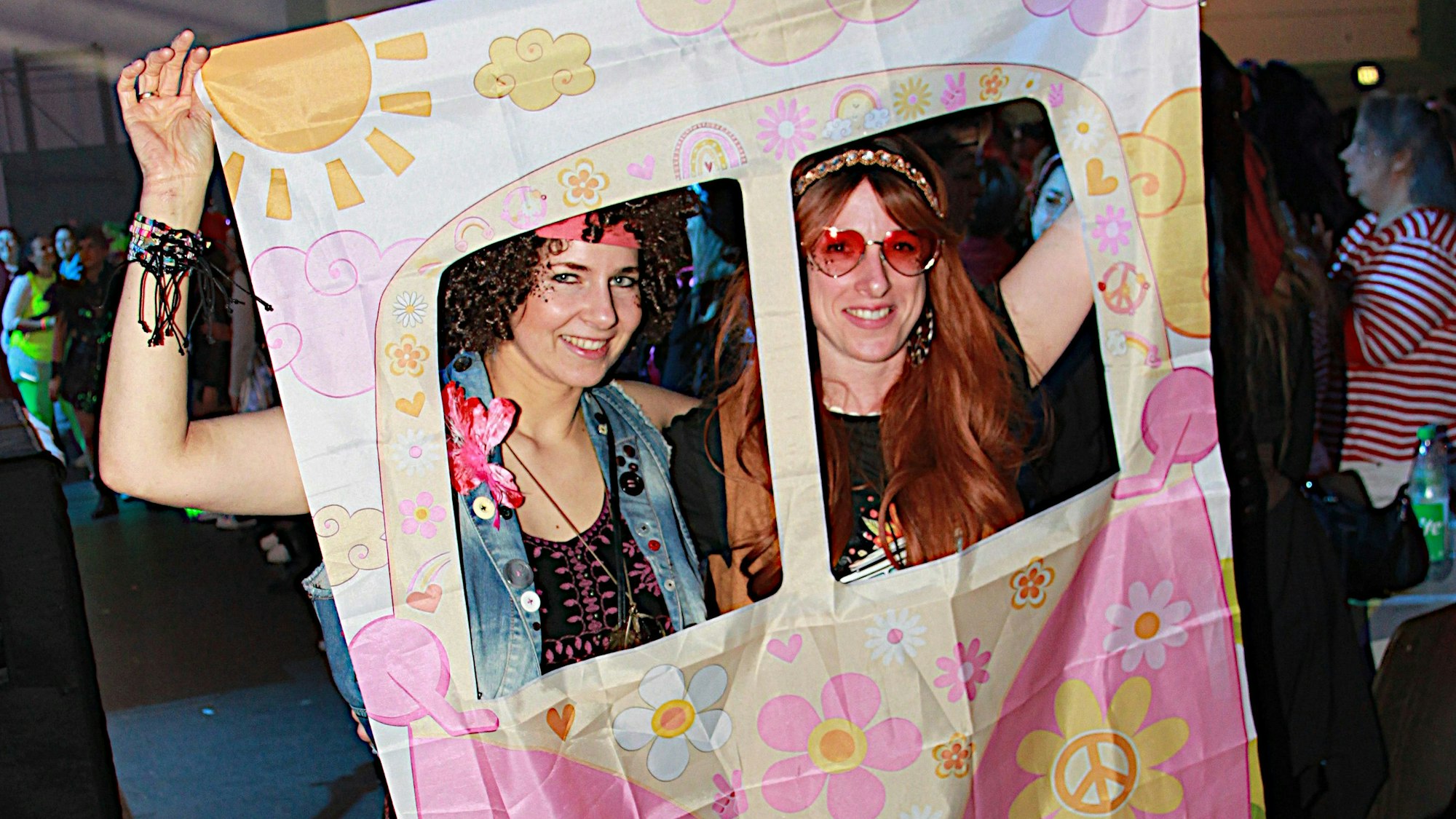 Zwei als Hippies verkleidete Frauen heben ein Tuch, das einen Bulli darstellen soll, und schauen aus den in Fensterform ausgeschnittenen Löchern.