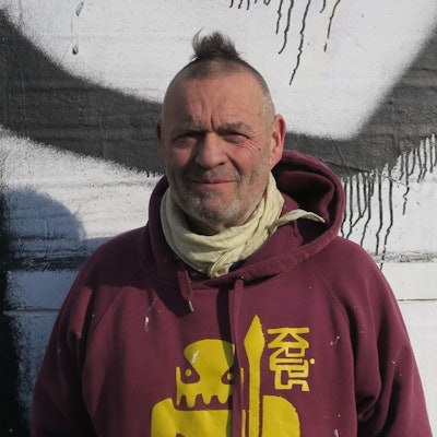 Ein Mann mit Mini-Iro im weinroten Pulle mit einem stilisierten Star-Wars-Kriege in Gelb darauf und dem Schriftzug KRIPS steht draußen vor einer besprühten Wand.