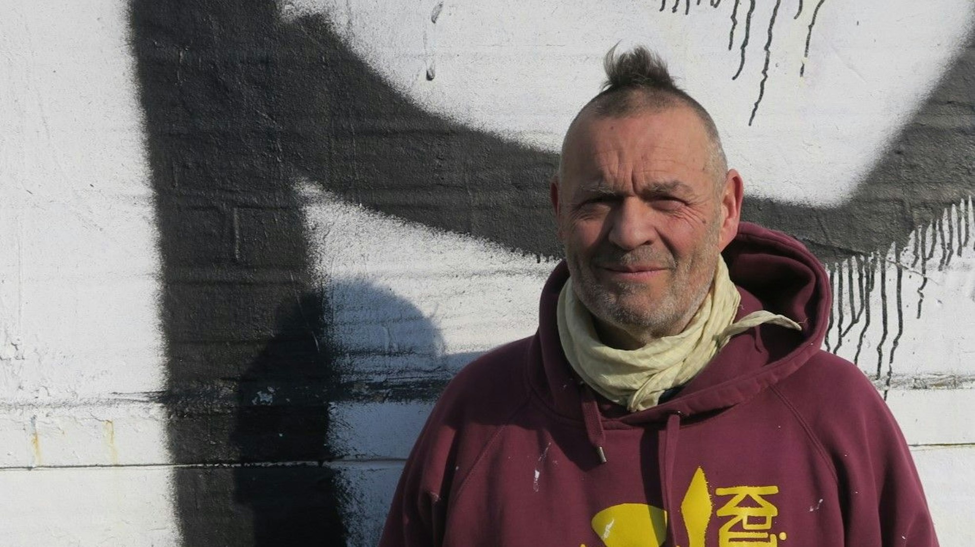 Ein Mann mit Mini-Iro im weinroten Pulle mit einem stilisierten Star-Wars-Kriege in Gelb darauf und dem Schriftzug KRIPS steht draußen vor einer besprühten Wand.