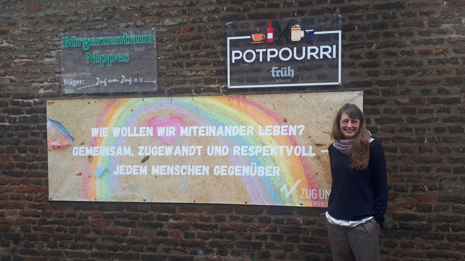 Eine Frau steht vor einer Mauer, neben ihr ist ein Schild zu sehen mit einem Regenbogen darauf.