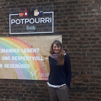 Eine Frau steht vor einer Mauer, neben ihr ist ein Schild zu sehen mit einem Regenbogen darauf.