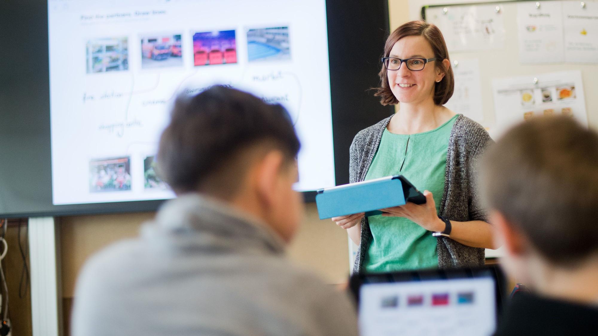 Eine Lehrerin steht mit einem iPad vor einer digitalen Schultafel im Englischunterricht einer 5. Klasse.