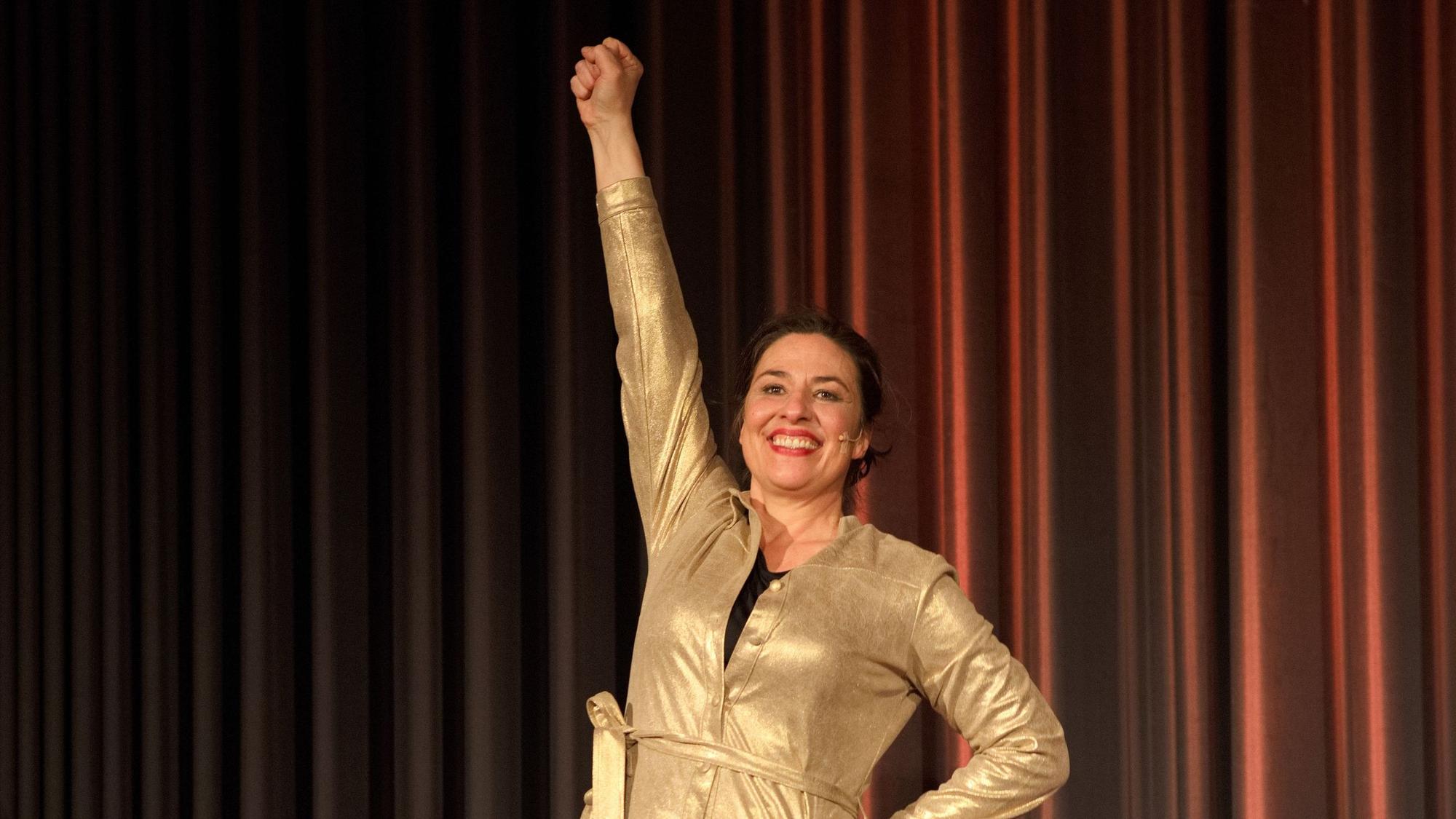 Die Kabarettistin Eva Eiselt steht in einem goldenem Kleid vor einem braunen Vorhang und reckt lachend die rechte Faust in die Luft.
