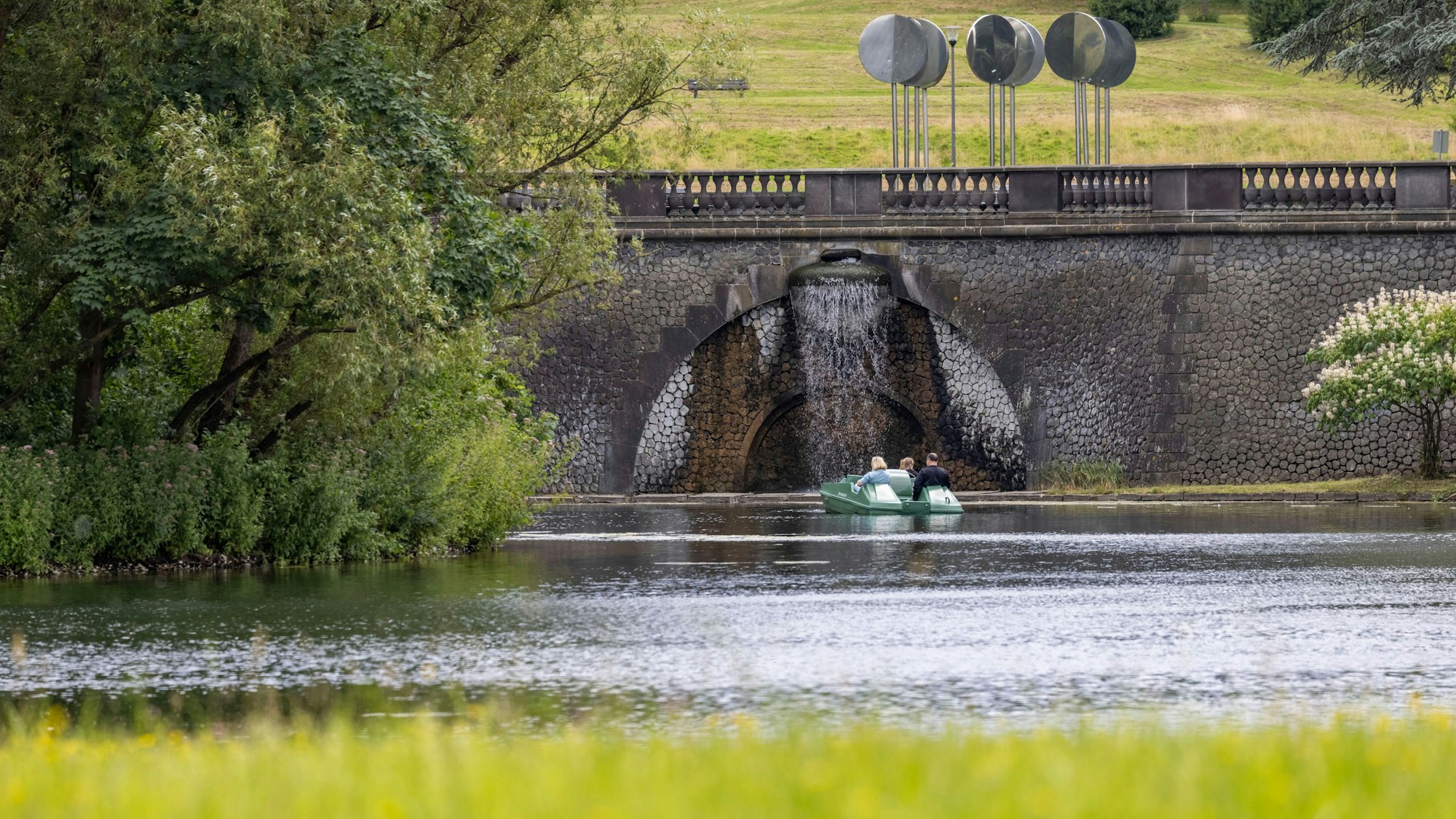 Ausflügler sind mit einem Tretboot bei sonnigem Wetter auf einem Teich in der Rheinaue unterwegs.