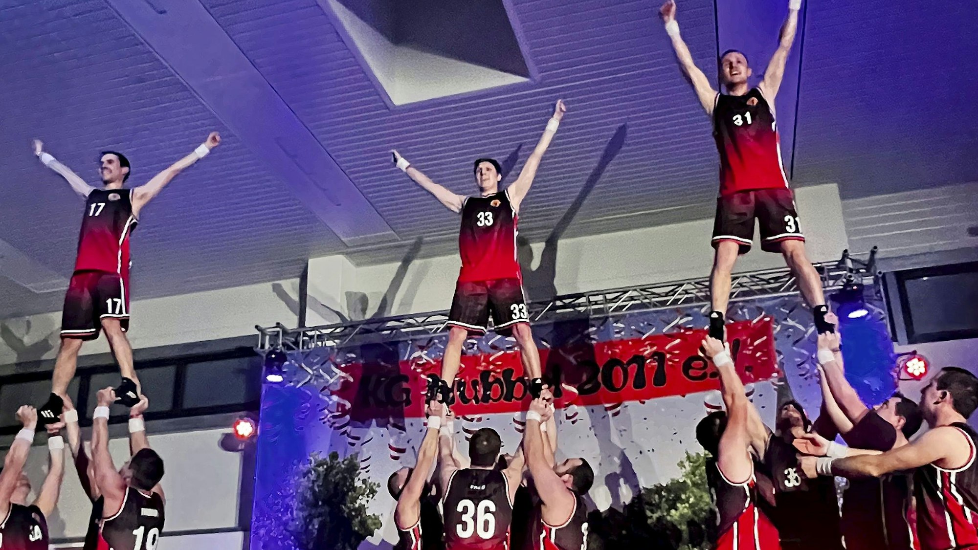 Eine Gruppe von Männern in schwarz-roten Sport-Outfits zeigt akrobatische Hebefiguren auf der Bühne.