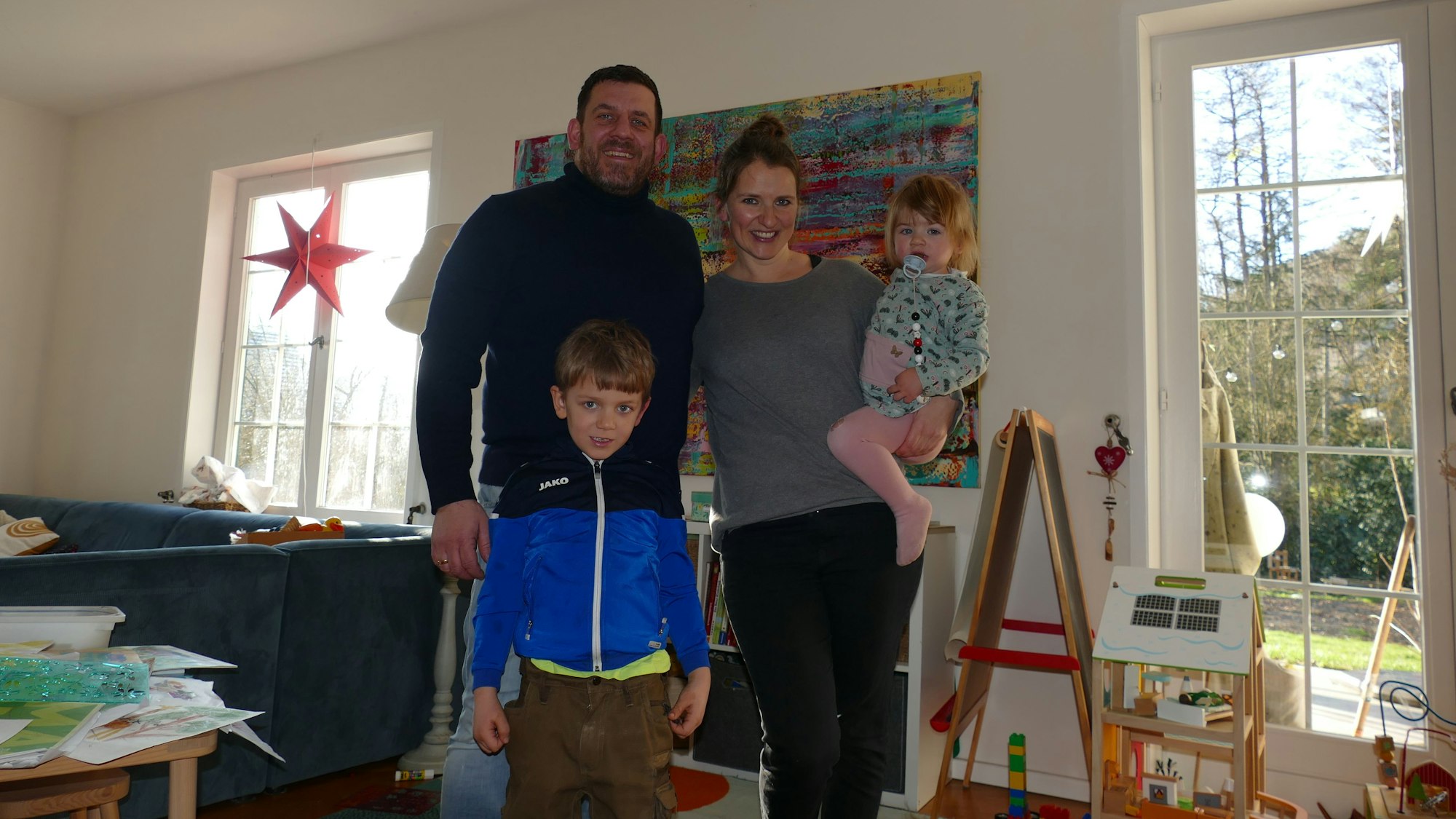 Eine vierköpfige Familie steht in einem Wohnzimmer mit großen Fenstern.