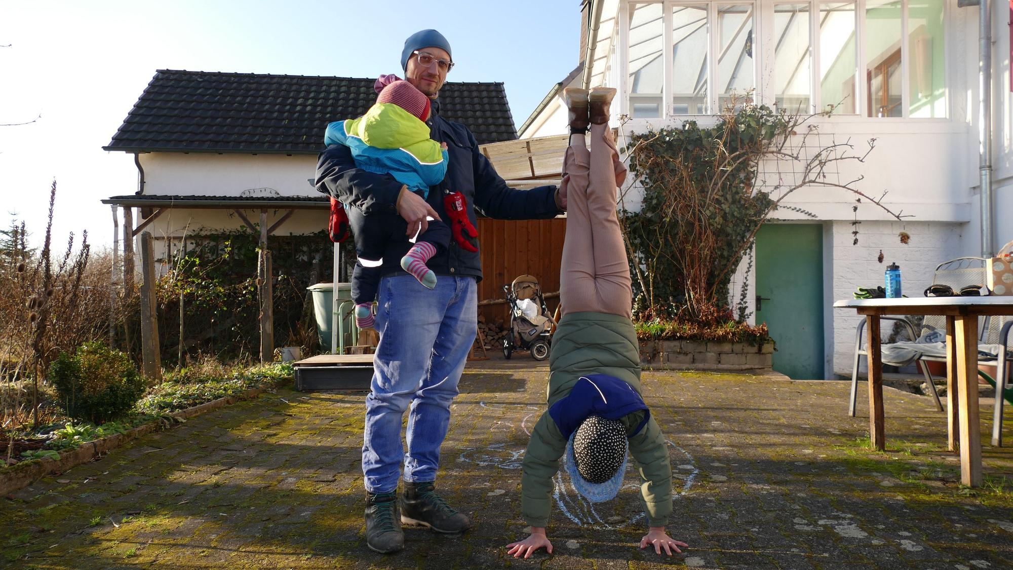 Ein Mann mit Mütze und Brille steht in einem Garten. Er hält ein Kind auf dem Arm, dessen Gesicht von der Kamera abgewandt ist. Ein weiteres Kind macht einen Handstand und schaut dabei in Richtung des Bodens.