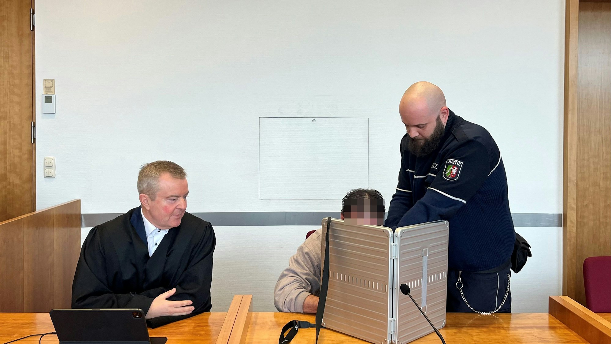 Der Angeklagte verbirgt sein Gesicht hinter einem Sichtschutz, neben ihm sitzt sein Verteidiger.