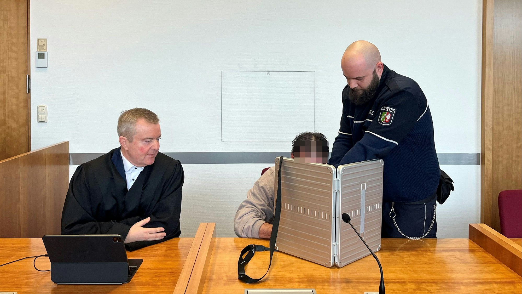 Der Angeklagte wird von einem Justizbediensteten in den Sitzungssaal des Bonner Landgerichts gebracht. Er verbirgt sein Gesicht hinter einem silberfarbenen schmalen Koffer, neben ihm sitzt sein Anwalt in schwarzer Robe.