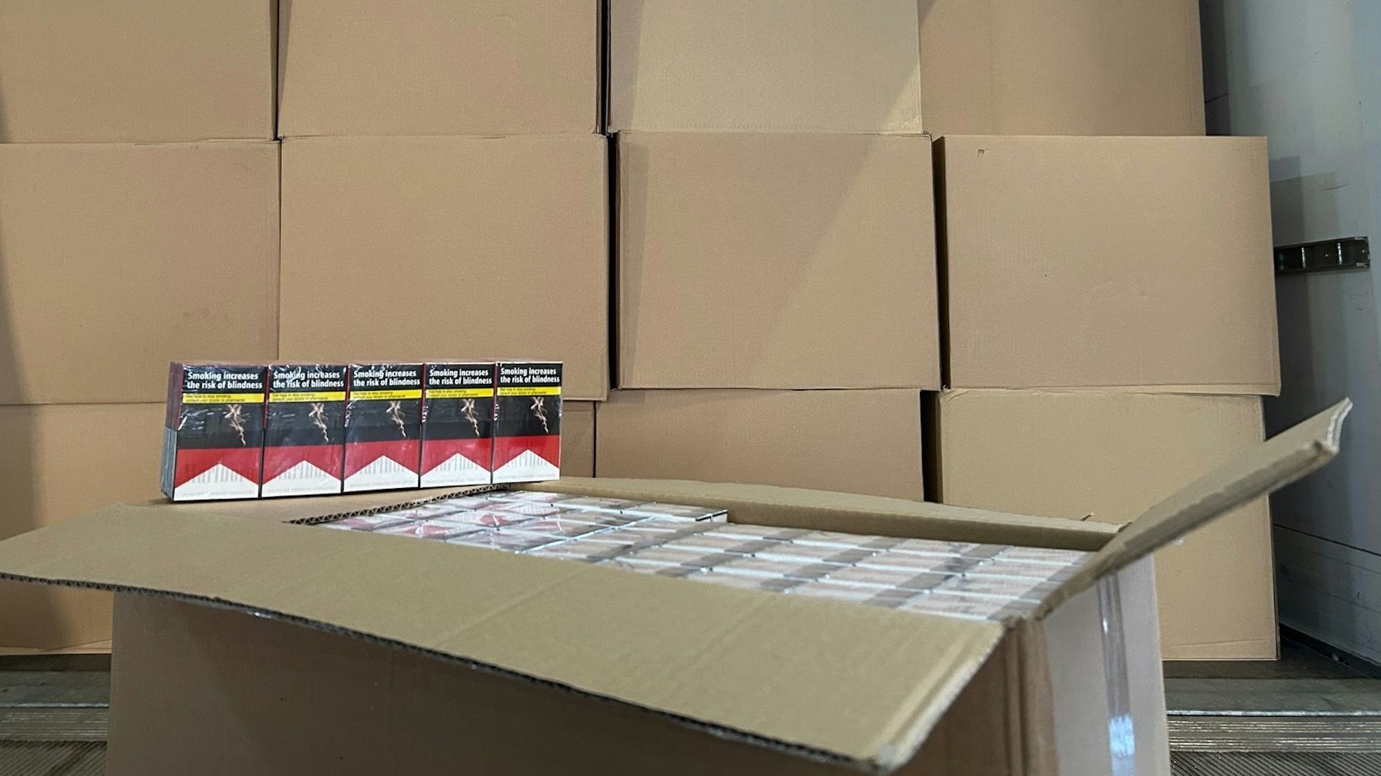 Eine geöffnete Pappkiste mit Zigarettenschachteln vor einer Wand aus geschlossenen Kartons.