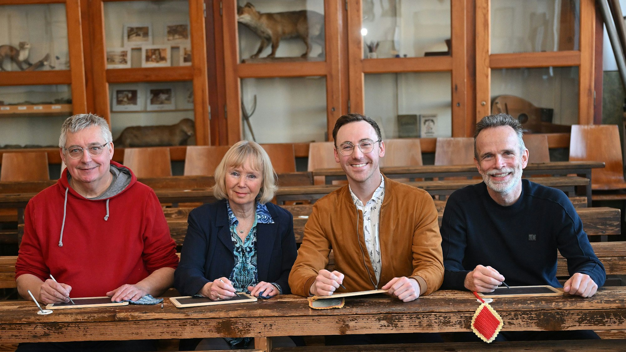 Drei Männer und eine Frau sitzen auf alten Schulbänken in einem Klassenraum.