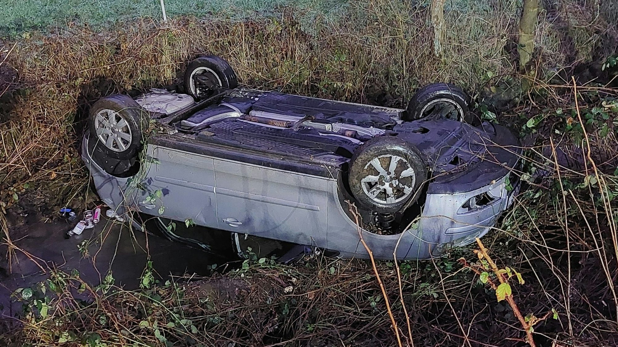 Ein Auto liegt auf dem Dach neben einer Straße über einem Bach.