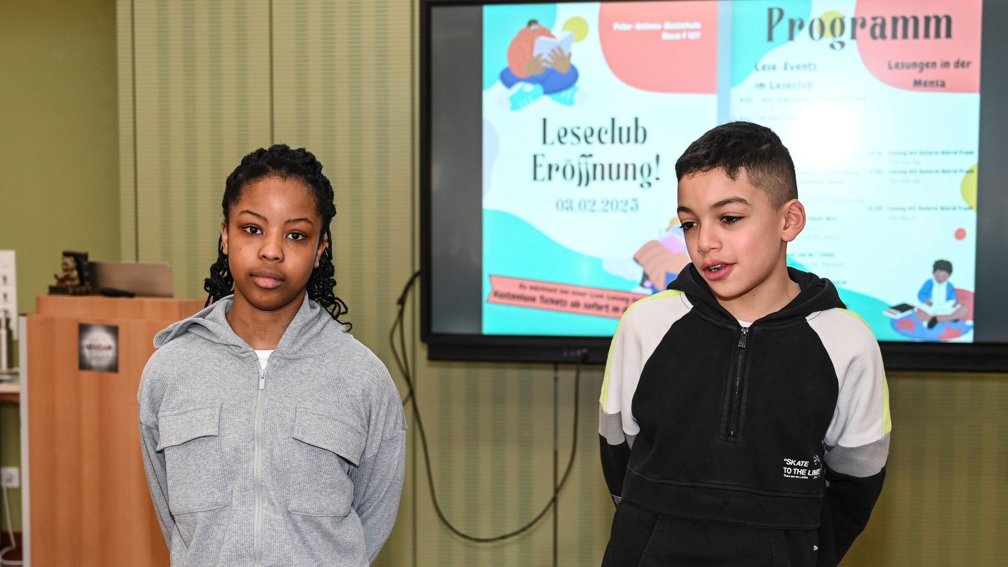 Eine Schülerin und ein Schüler der Peter-Ustinov-Realschule stehen vor dem Monitor, auf dem das Programm der Eröffnungsfeier des Leseclubs zu sehen ist.