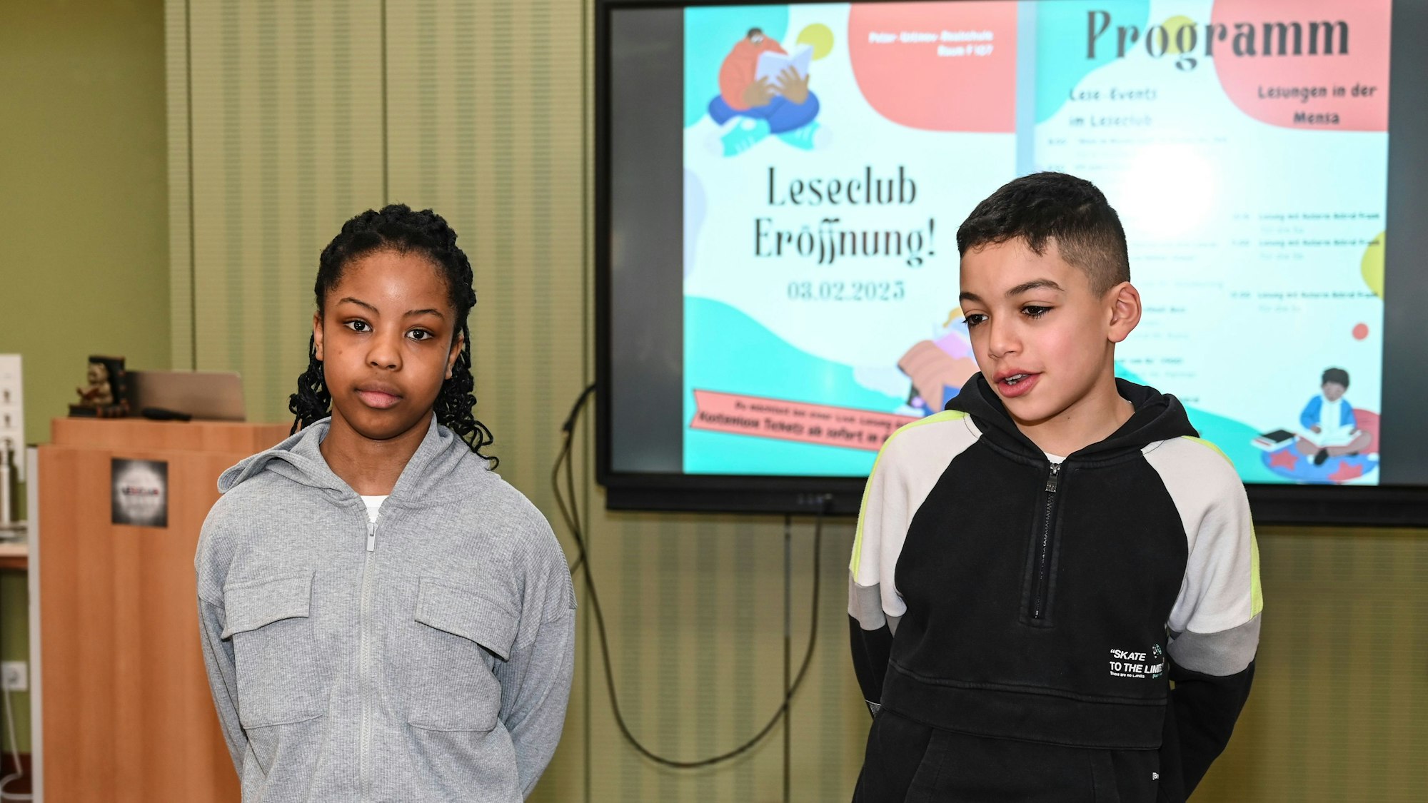 Eine Schülerin und ein Schüler der Peter-Ustinov-Realschule stehen vor dem Monitor, auf dem das Programm der Eröffnungsfeier des Leseclubs zu sehen ist.