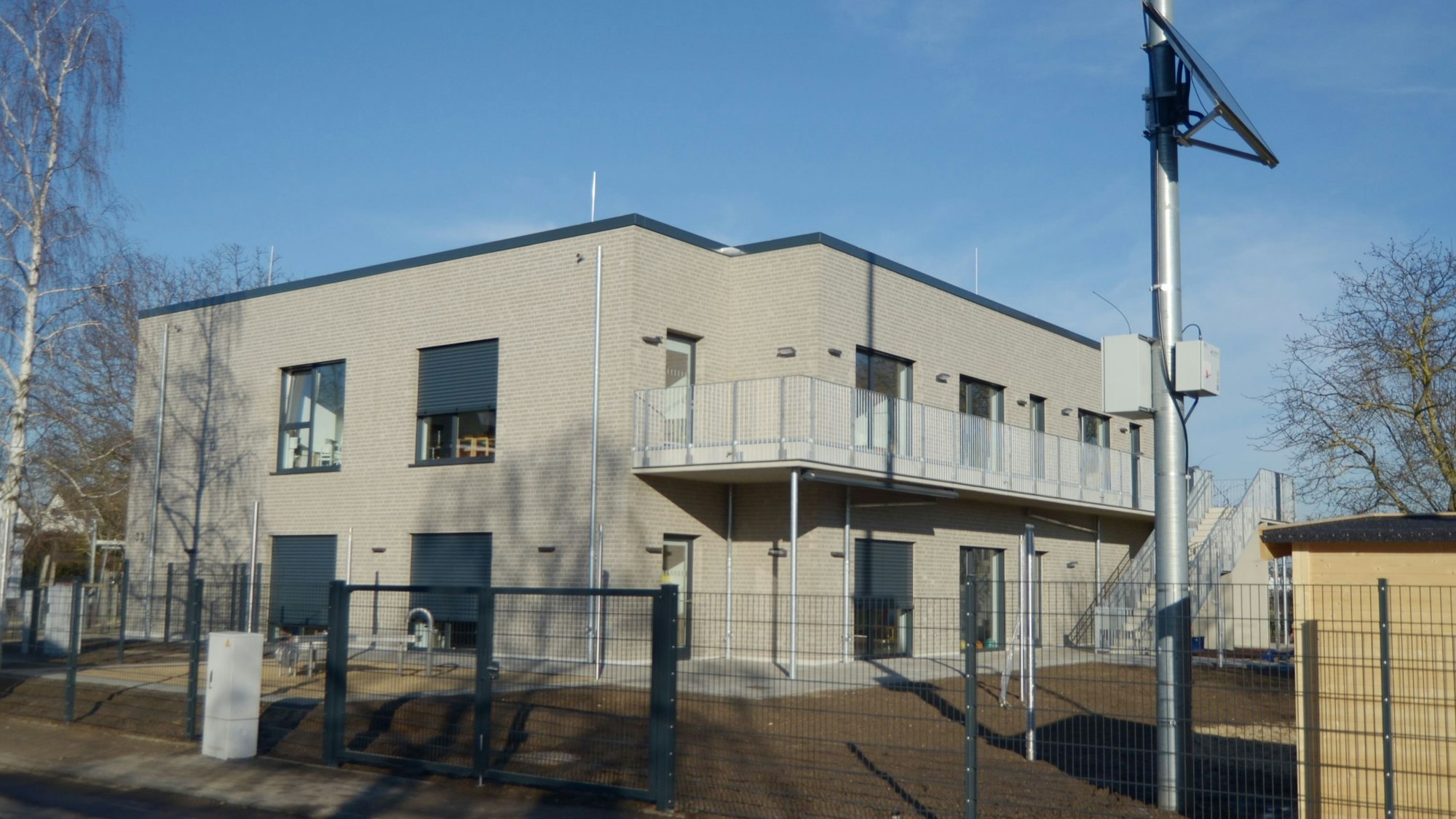 Der zweigeschossige Bau mit Balkon ist grau verklinkert, das Außengelände von einem Zaun umgeben.