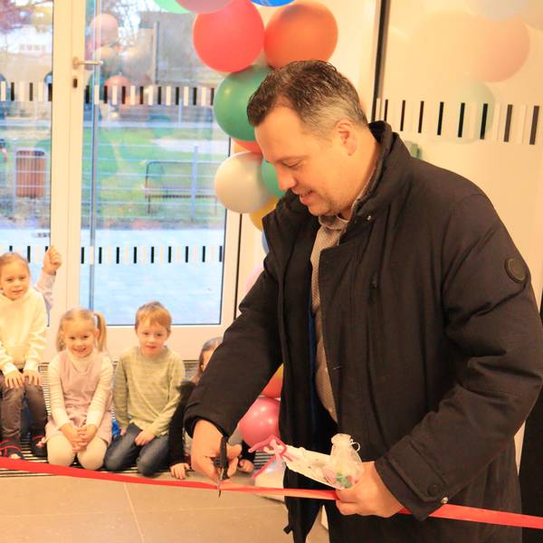 Sacha Reichelt schneidet ein rotes Band durch. Mehrere Kinder und eine Erzieherin im Hintergrund schauen zu.