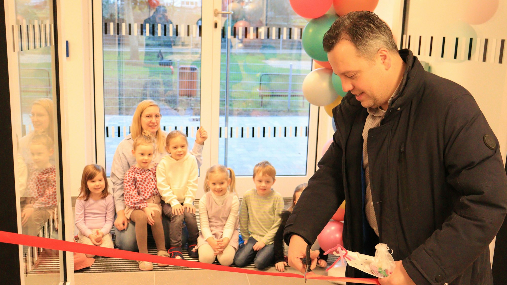 Sacha Reichelt schneidet ein rotes Band durch. Mehrere Kinder und eine Erzieherin im Hintergrund schauen zu.