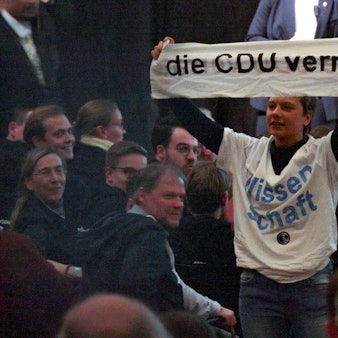 Eine Aktivistin mit einem T-Shirt mit der Aufschrift „Wissenschaft“ hält während einer Rede von Unionskanzlerkandidat Merz auf einer Wahlkampfveranstaltung im WCCB ein Transparent mit dem Satz „Die CDU verrät“ hoch.