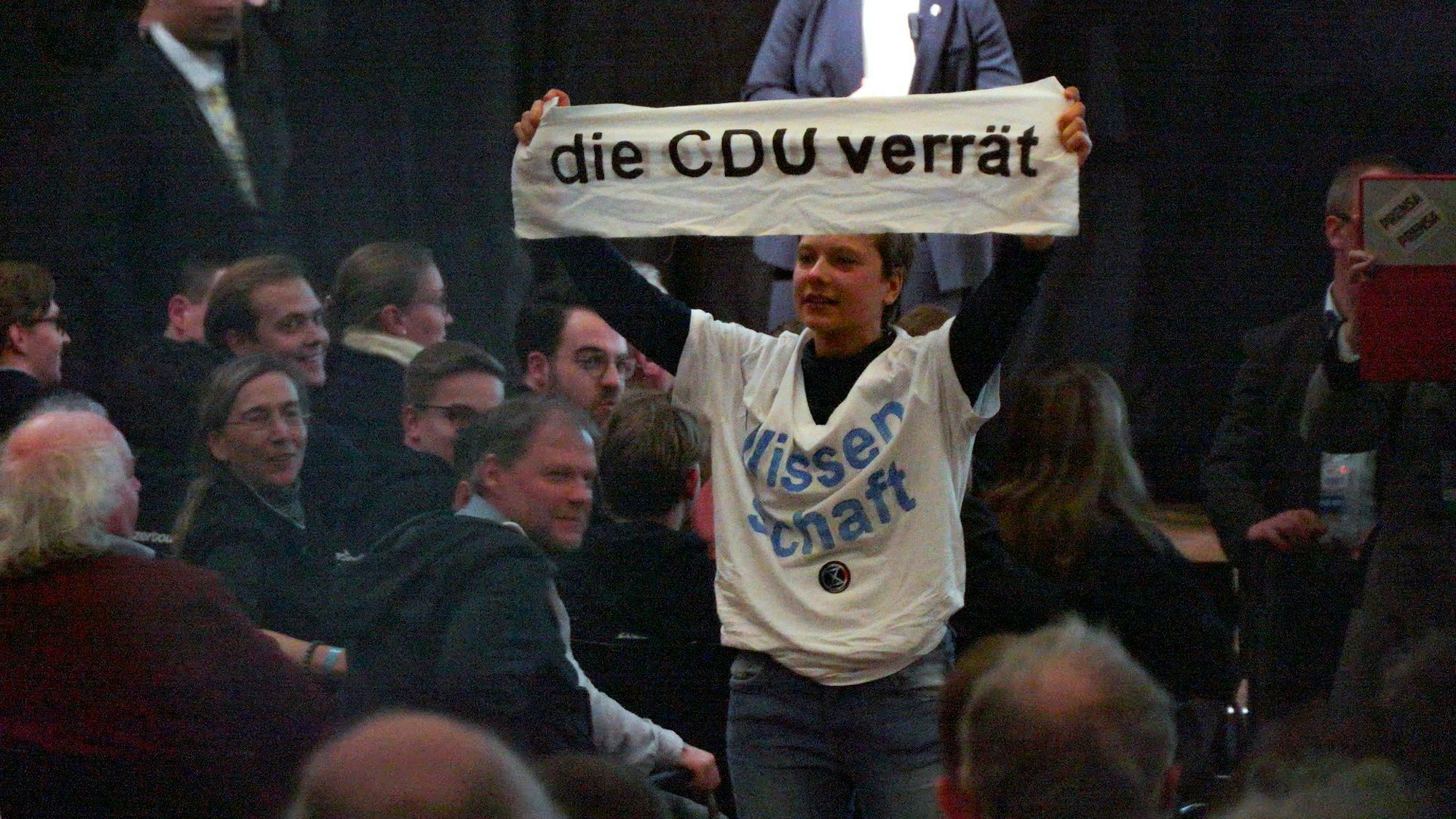 Eine Aktivistin mit einem T-Shirt mit der Aufschrift „Wissenschaft“ hält während einer Rede von Unionskanzlerkandidat Merz auf einer Wahlkampfveranstaltung im WCCB ein Transparent mit dem Satz „Die CDU verrät“ hoch.