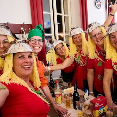 Eine Gruppe Frauen hat sich lustige Kostüme gebastelt: Zum tomatenroten Shirt tragen sie Nudelsiebe auf dem Kopf, die daran befestigten Fäden sehen aus wie Spaghetti.