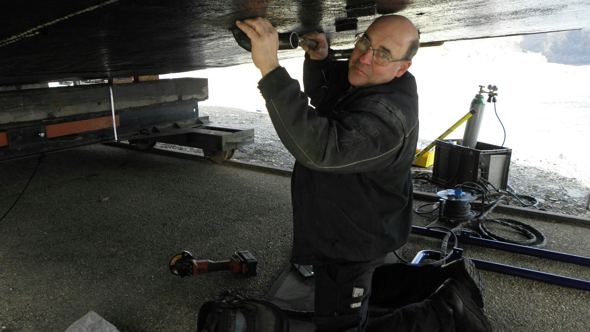 Ein Mann in Arbeitskleidung kniet auf dem Boden und arbeitet an einem Rohr an der Unterseite eines Schiffes, das aus dem Rursee an Land gezogen wurde.