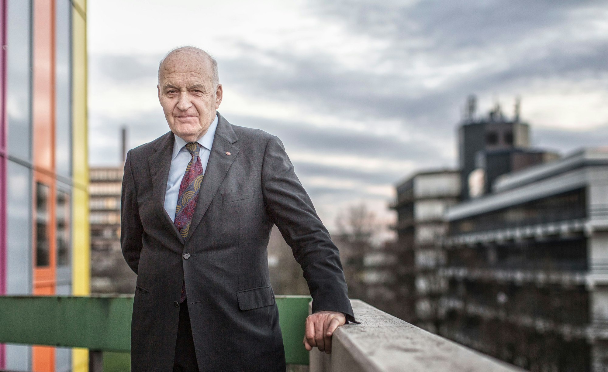 Der 2022 verstorbene DM-Gründer Götz Werner ist im Halbporträt zu sehen auf einem Balkon vor großstädtischer Kulisse.