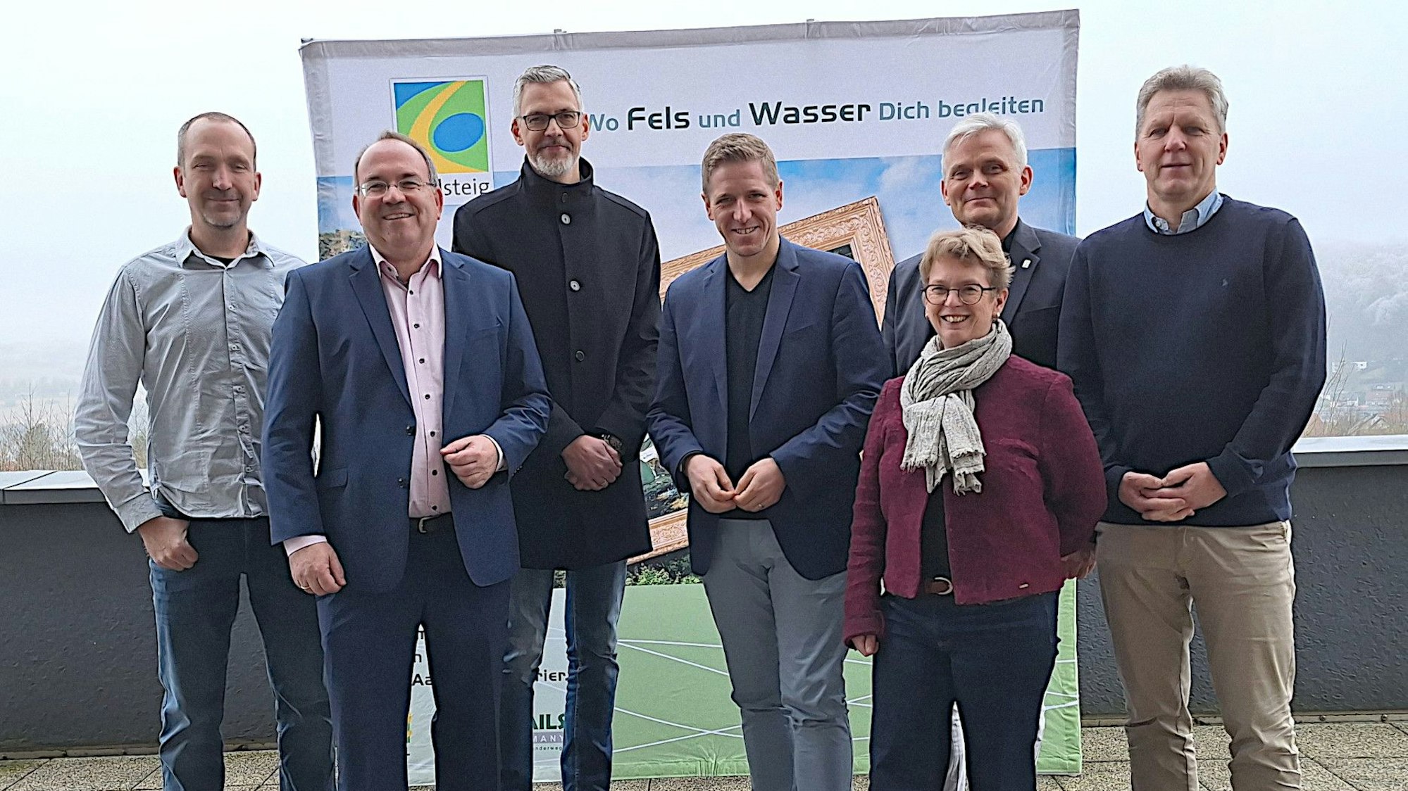 Sechs Männer und eine Frau stehen vor einem Plakat, das für den Eifelsteig wirbt.