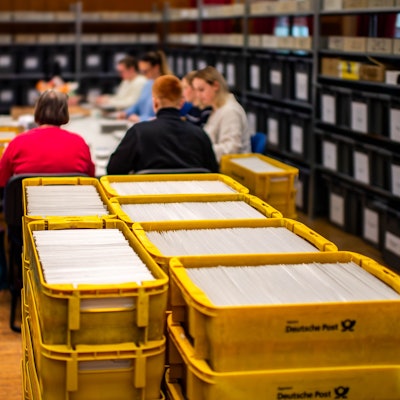Das Bild zeigt zahlreiche gelbe Postkisten im Vordergrund, die randvoll mit Wahlunterlagen sind.