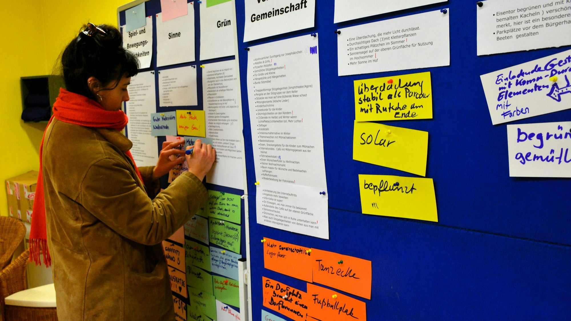 Eine Frau mit brauner Jacke und rotem Schal steht vor einer blauen Notizwand auf der viele teils bunte Zettel stecken.