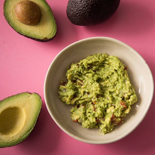 Zerdrückte und gewürzte Avocado als Brotaufstrich in einer Schüssel. Daneben offene Avocadohälften.