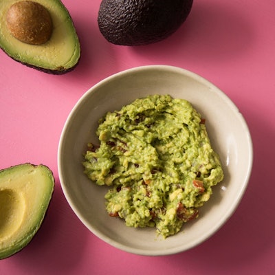 Zerdrückte und gewürzte Avocado als Brotaufstrich in einer Schüssel. Daneben offene Avocadohälften.