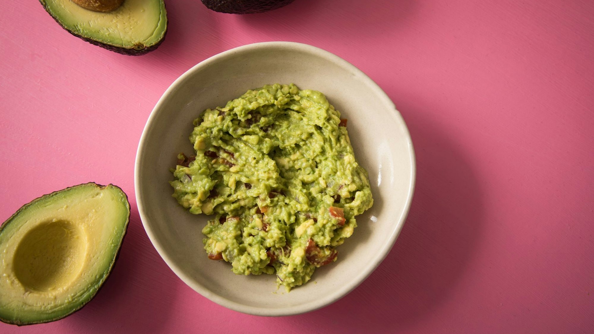 Zerdrückte und gewürzte Avocado als Brotaufstrich in einer Schüssel. Daneben offene Avocadohälften.