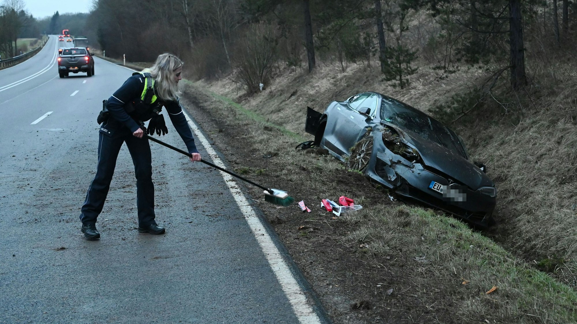 Das Bild zeigt eine Polizistin, die mit einem Besen Schrott in den Straßengraben kehrt. In dem liegt bereits der Tesla.