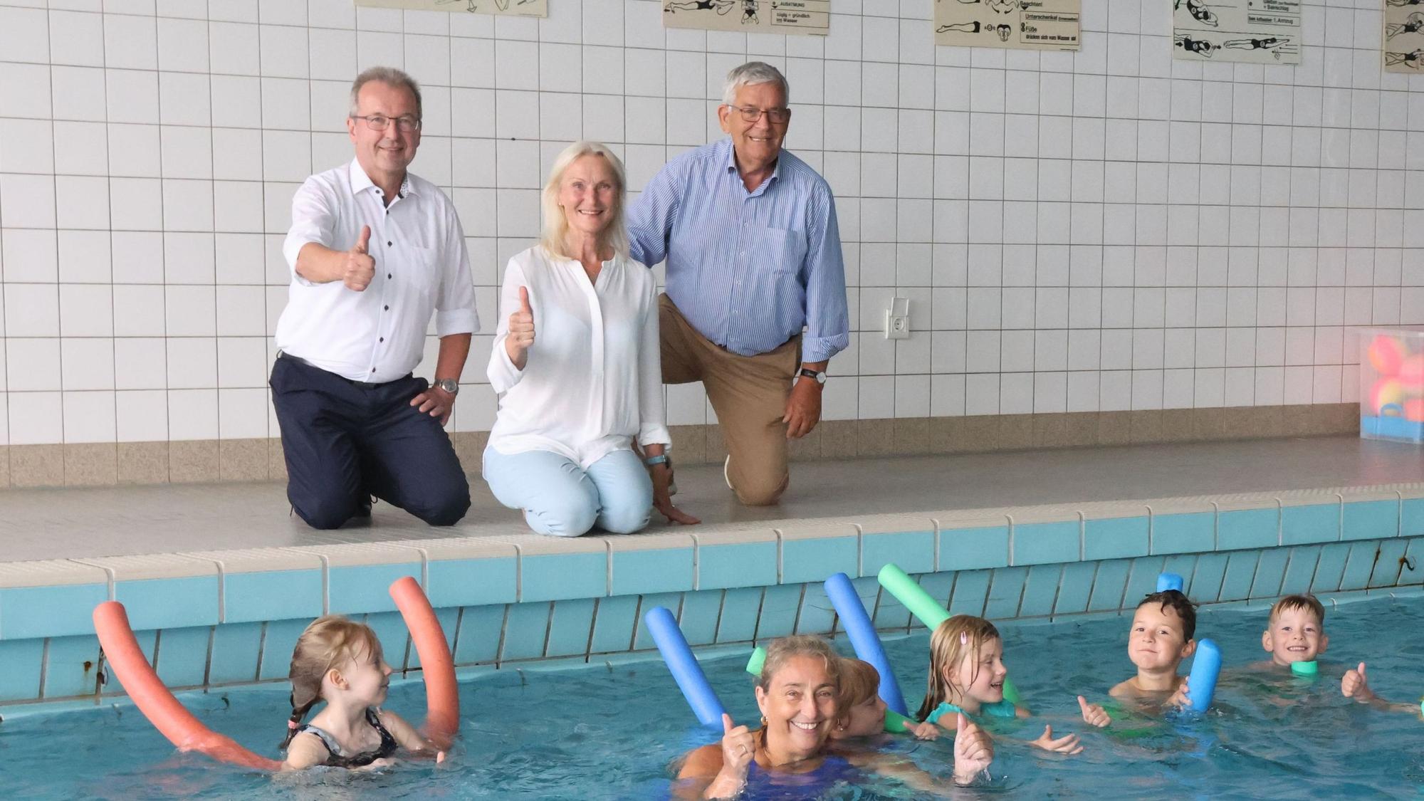 Eine Frau und zwei Männer am Rand eines Schwimmbeckens, darin einige Kinder.