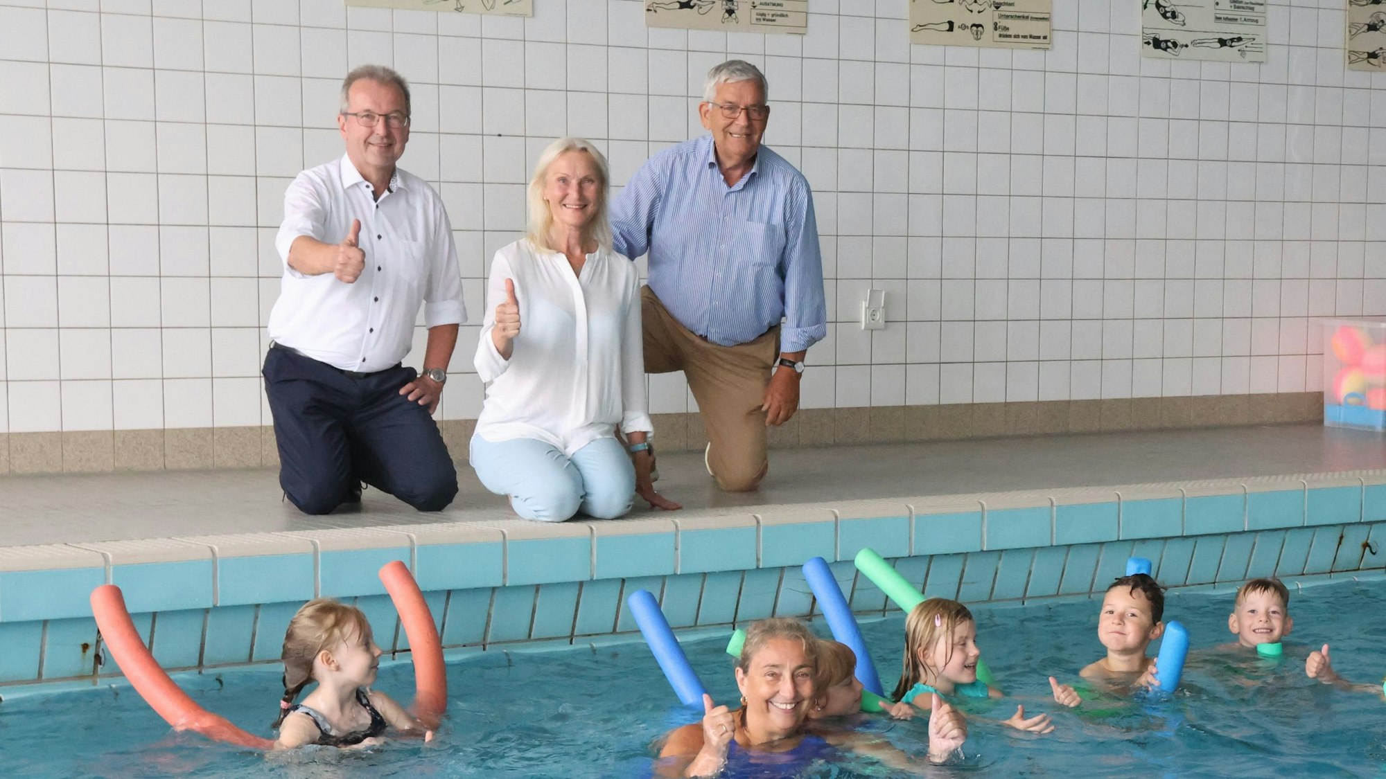 Eine Frau und zwei Männer am Rand eines Schwimmbeckens, darin einige Kinder.