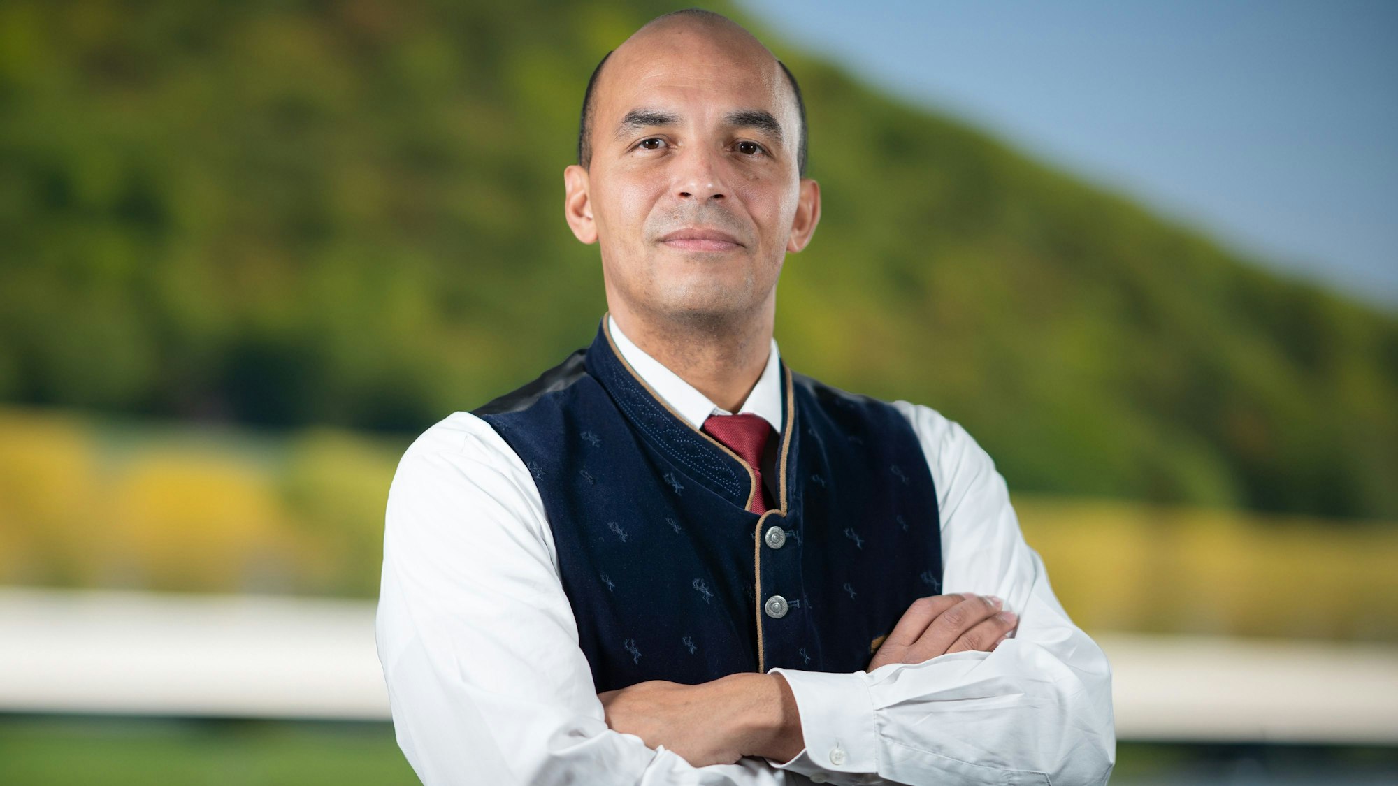 Ein Mann in einer Trachtenweste, roter Krawatte und weißem Hemd steht mit verschränkten Armen vor einem verschwommenen grünen Hintergrund.