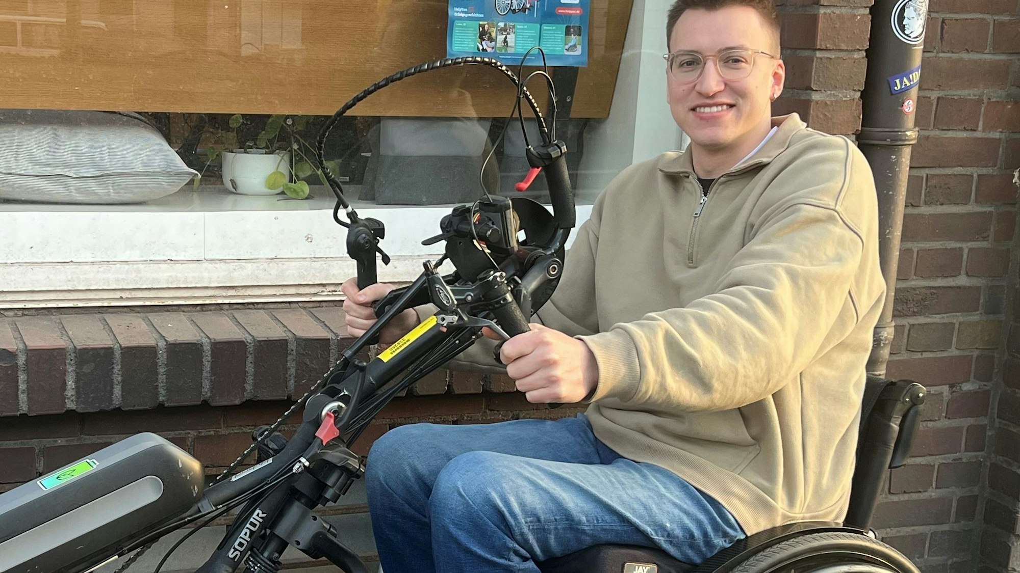 Ein junger Mann sitzt auf einem Handbike. Er steht damit auf dem Bürgersteig, im Hintergrund ist ein Fenster.