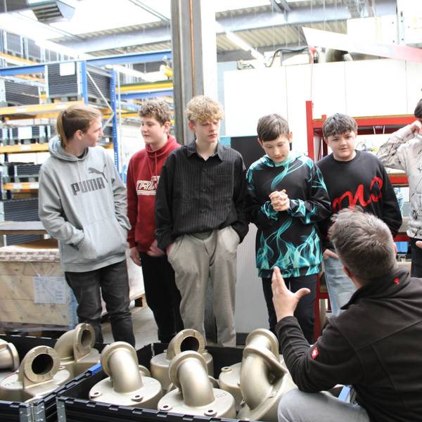 Sieben Jungen stehen in einer Fabrikhalle, ein Mitarbeiter hockt gestikulierend vor ihnen.