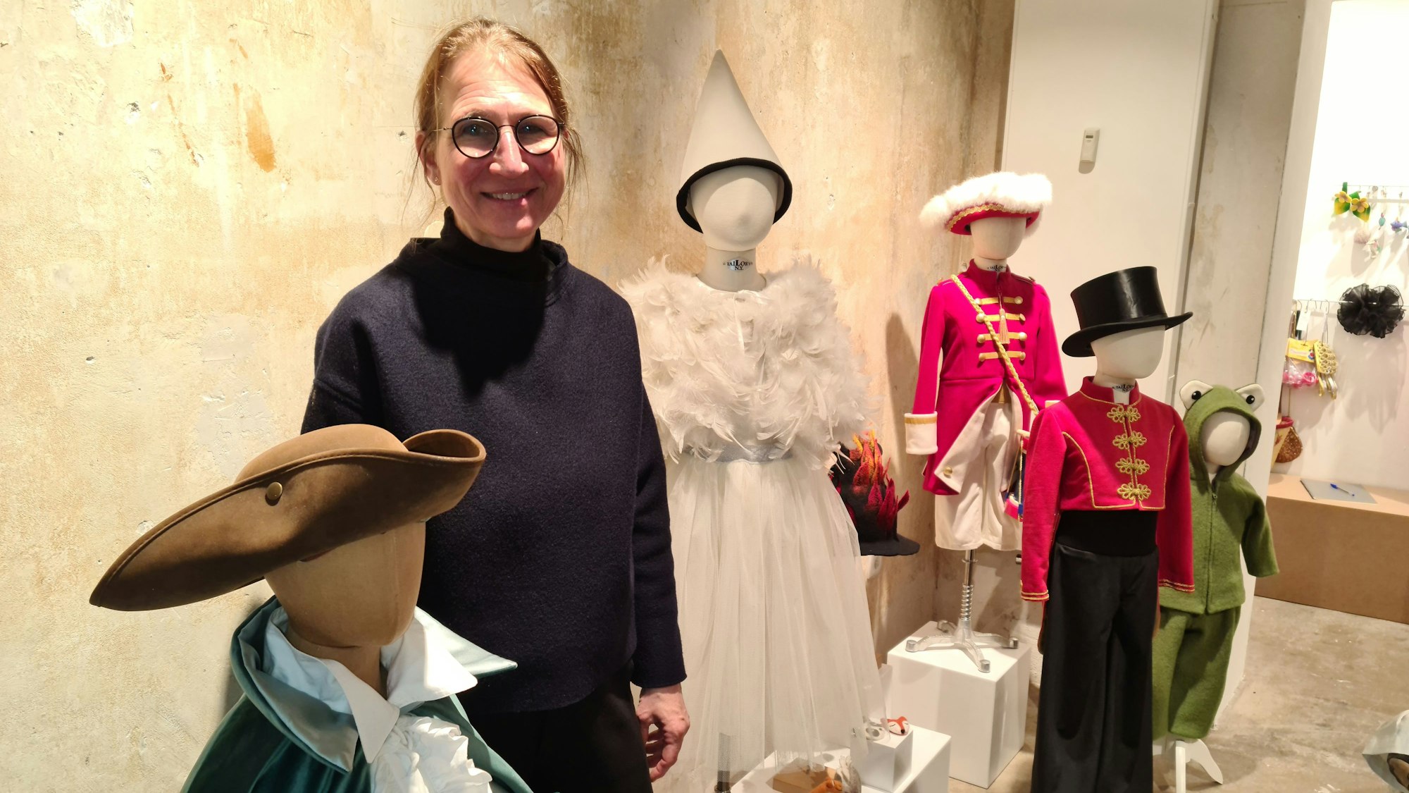 Eine Frau mit Brille steht neben mehreren Schaufensterpuppen in Kindergröße, die Kostüme oder ein Kleidchen anhaben.