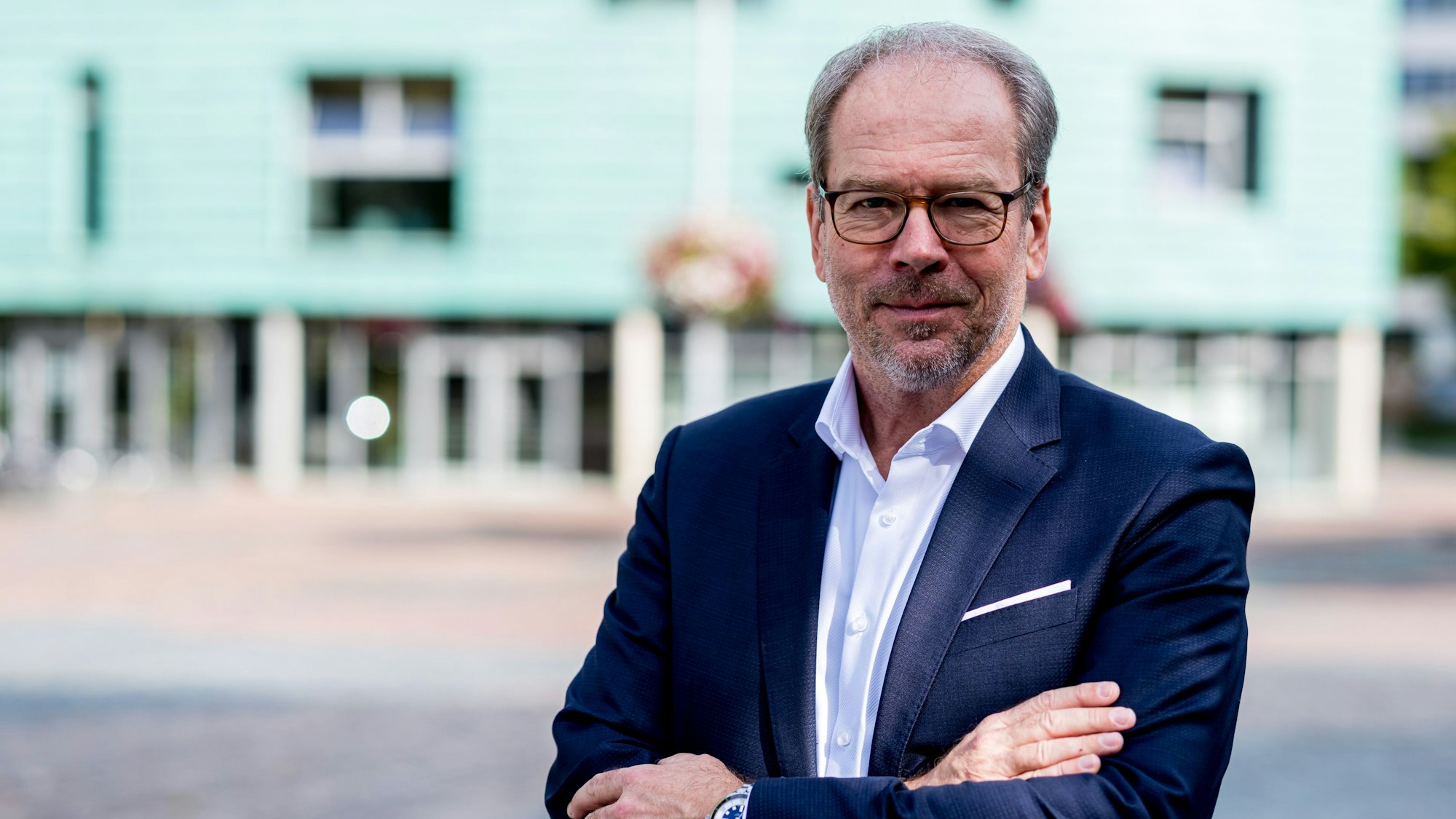 Der Bürgermeister trägt einen dunkelblauen Anzug und hat die Arme verschränkt. Im Hintergrund verschwommen die Stadtbibliothek.