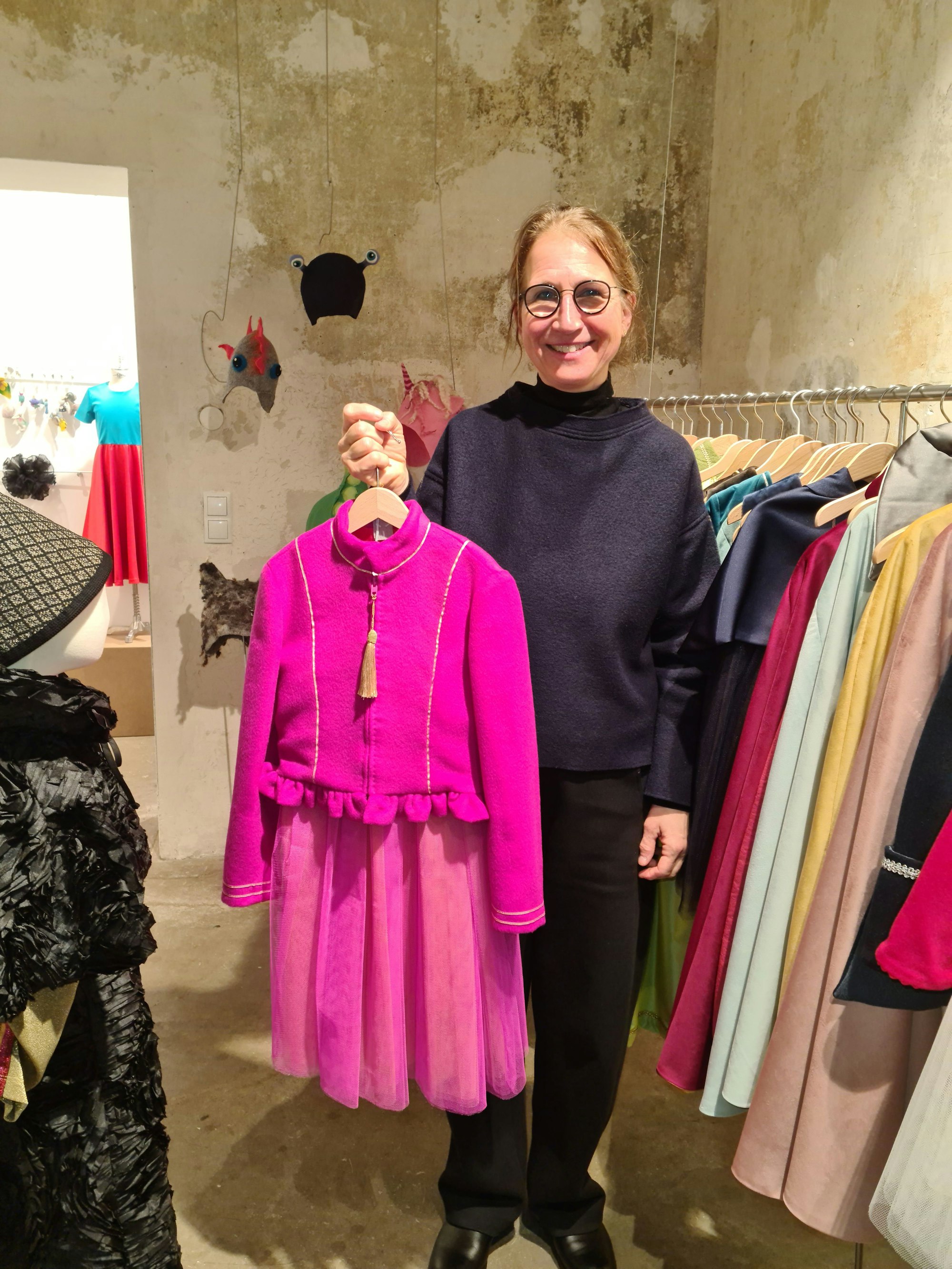 Eine Frau mit Brille hält ein pinkfarbenes Kleid in die Höhe, hinter ist ihr ist eine Kleiderstange mit weiteren Kleidungsstücken zu sehen.