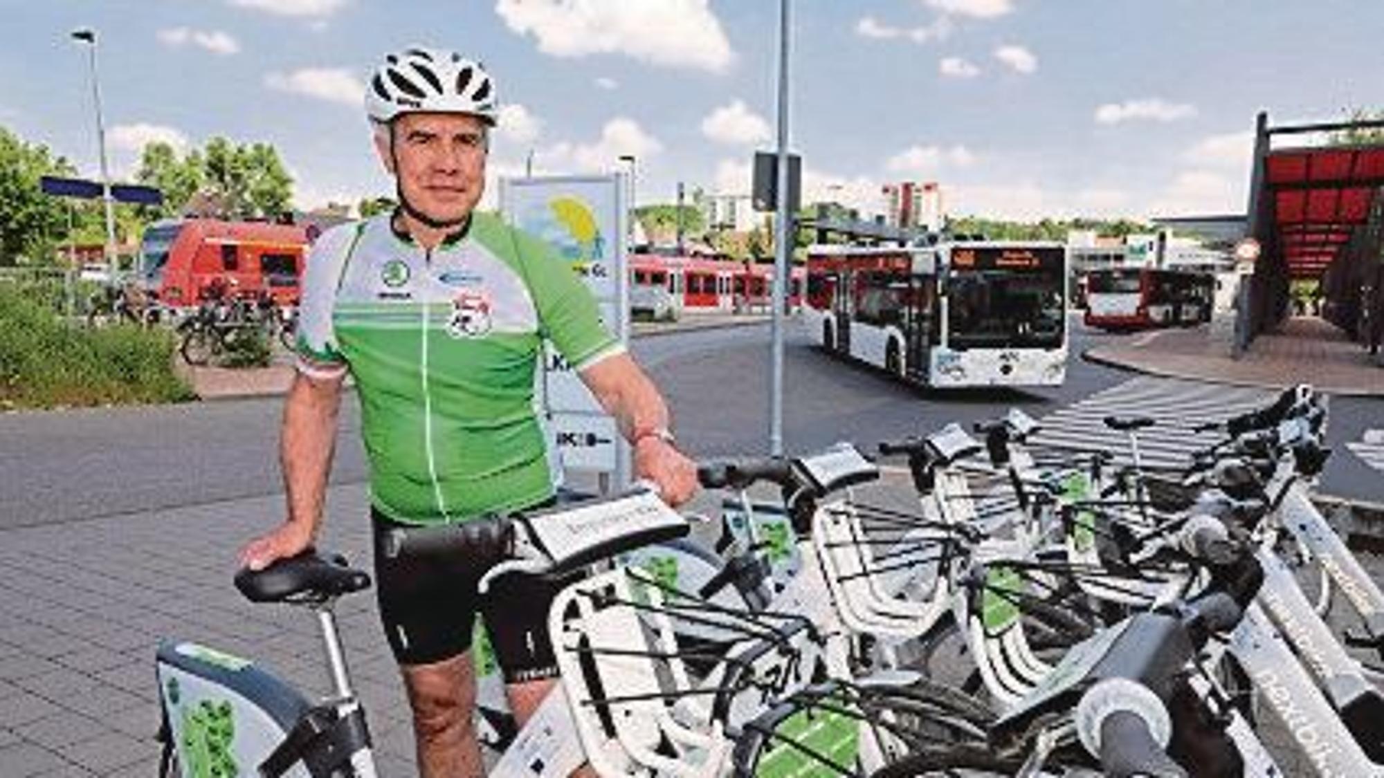 Ein Fahrradfahrer steht vor einer Reihe Bergischer E-Bikes. Im Hintergrund sind eine Bahnstation und ein Bus zu sehen.