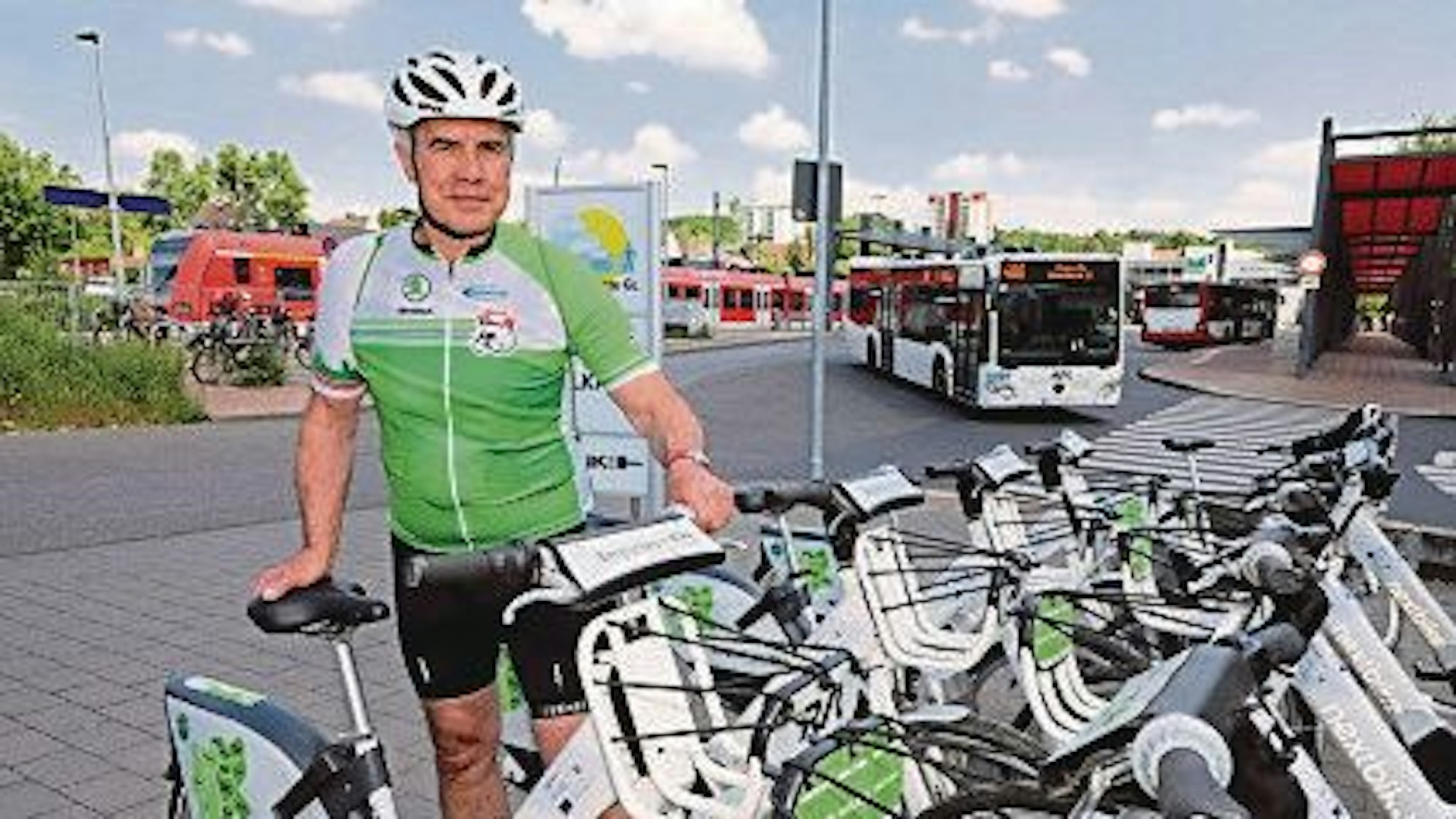 Ein Fahrradfahrer steht vor einer Reihe Bergischer E-Bikes. Im Hintergrund sind eine Bahnstation und ein Bus zu sehen.