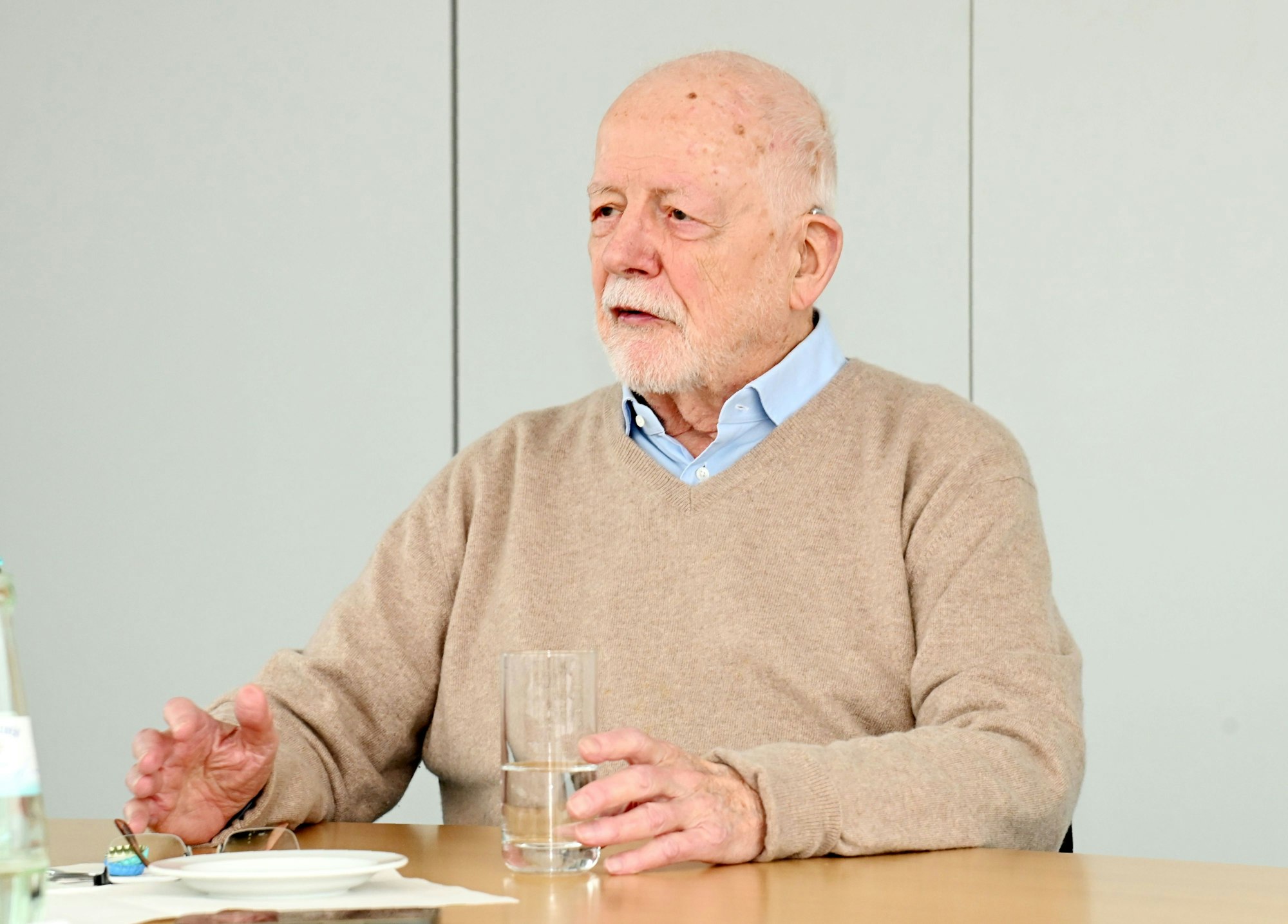 Der Politikwissenschaftler ist in einer Gesprächssituation zu sehen. Er trägt einen beigen Pullover und ein hellblaues Hemd. Vor ihm steht ein Glas Wasser.