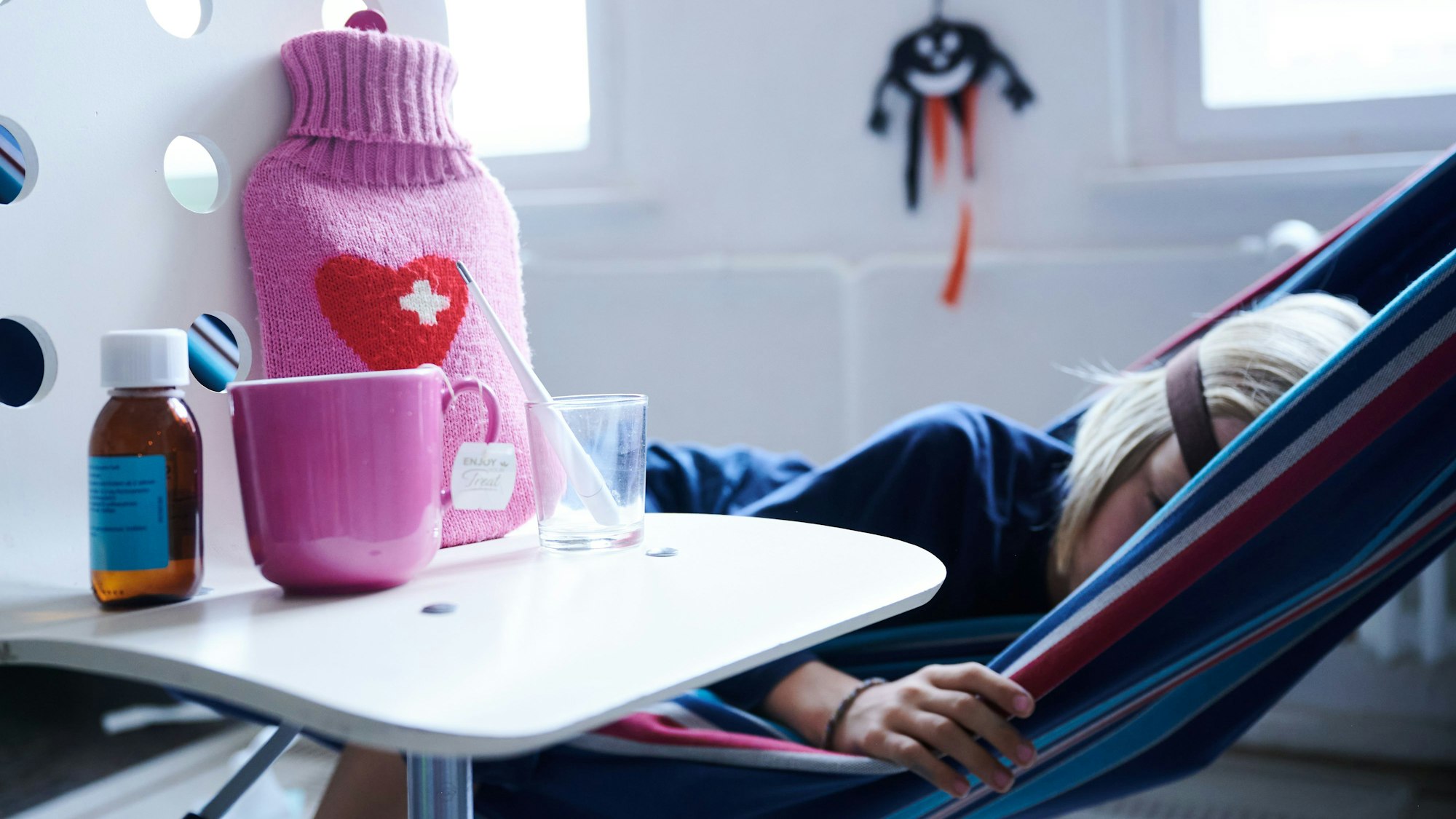 Ein Kind liegt in der Hängematte, daneben Wärmflasche und Medizin.