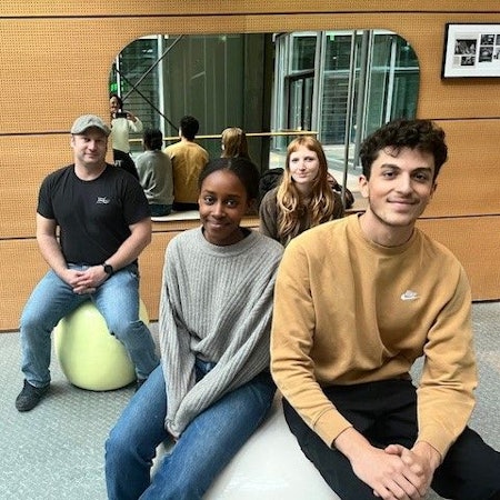 Eine vier-köpfige Delegation der Bezirksschüler*innenvertetung sitzt auf runden Plastiksitzen im Eingangsbereich des DuMont-Verlagshauses vor einem großen Spiegel.