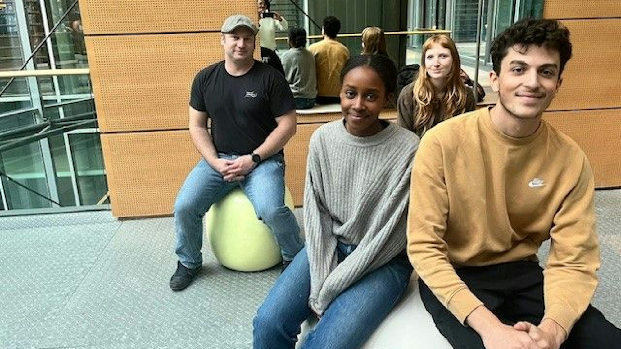 Eine vier-köpfige Delegation der Bezirksschüler*innenvertetung sitzt auf runden Plastiksitzen im Eingangsbereich des DuMont-Verlagshauses vor einem großen Spiegel.