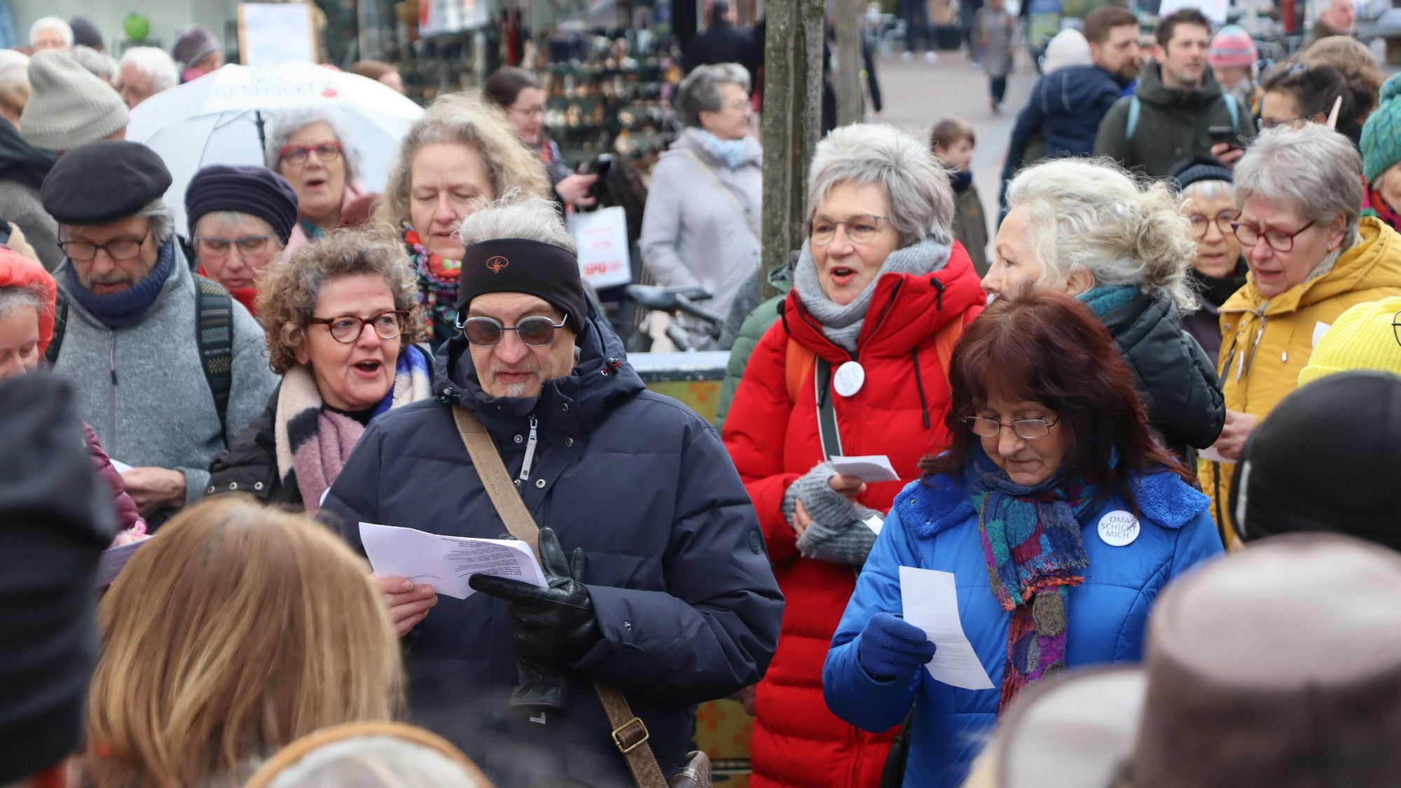 Auf dem Bild ist eine Menschenmenge in der Brühler Fußgängerzone zu sehen. Die Leute halten Zettel mit Liedtexten in der Hand und singen.